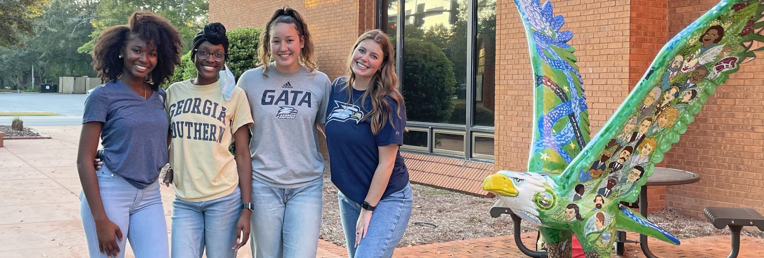 Four female students smiling and posing together outdoors next to a colorful, painted eagle sculpture in front of a brick campus building