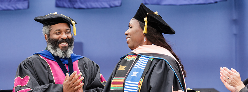 A smiling male faculty member with a full grey beard in doctoral regalia applauding a female graduate wearing a black tam, kente cloth stole, and academic gown.