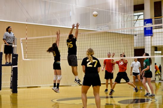 An indoor volleyball game in progress inside a gymnasium, featuring players jumping near the net to block/hit the ball while an official oversees from the referee stand.