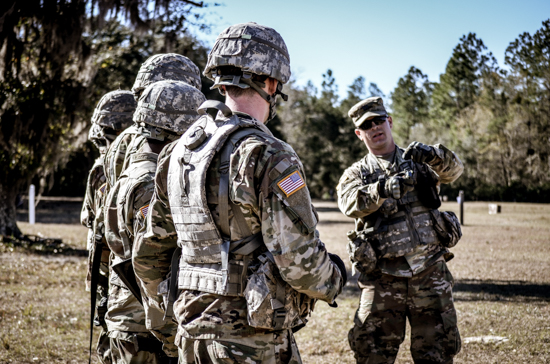 An Army ROTC instructor in tactical gear addressing a line of cadets wearing helmets and combat uniforms during an outdoor field training exercise.