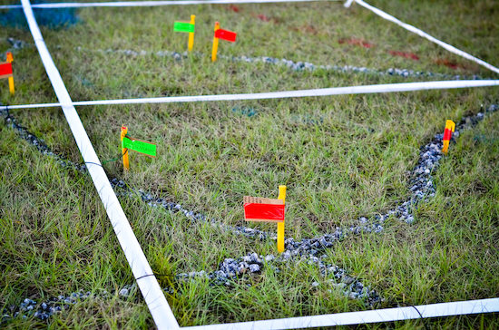 A close-up outdoor photograph of a gridded field training area marked by white tape, featuring small green and red flags placed along trails of crushed gray rock on a grassy surface.