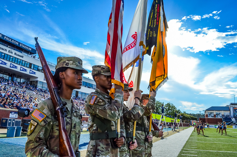 An Army ROTC color guard standing at attention with rifles and flags on the football field inside Paulson Stadium during a game day event.