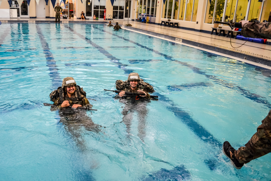 Two Army ROTC cadets in full combat uniform, helmets, and rucksacks treading water while holding training rifles during combat water survival testing in an indoor swimming pool.