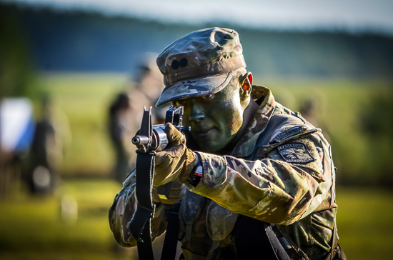 An Army ROTC cadet with face paint wearing camouflage tactical gear aiming a rifle directly toward the camera during an outdoor field training exercise