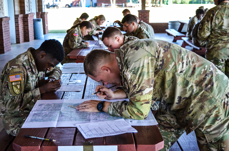Army ROTC cadets in camouflage uniforms sitting at wooden picnic tables under an outdoor pavilion studying paper maps during a land navigation training session.