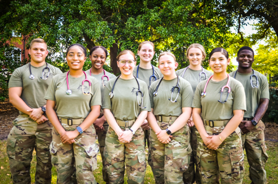 A group portrait of eight smiling military nursing/medical students or ROTC cadets standing outdoors in military physical training/combat uniforms with stethoscopes around their necks.