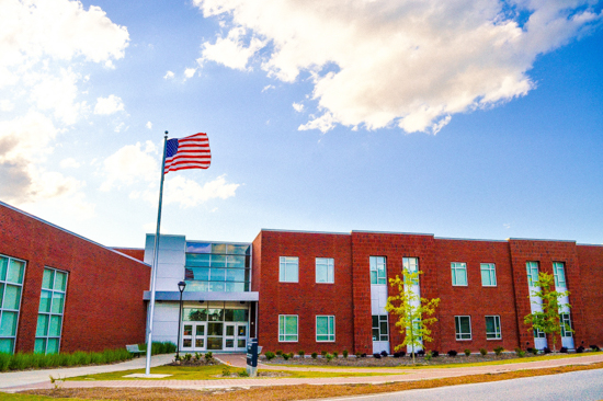 A two-story red brick modern campus building with large glass entry doors, silver accent panels, young trees out front, and an American flag flying high on a flagpole to the left under a partly cloudy sky.