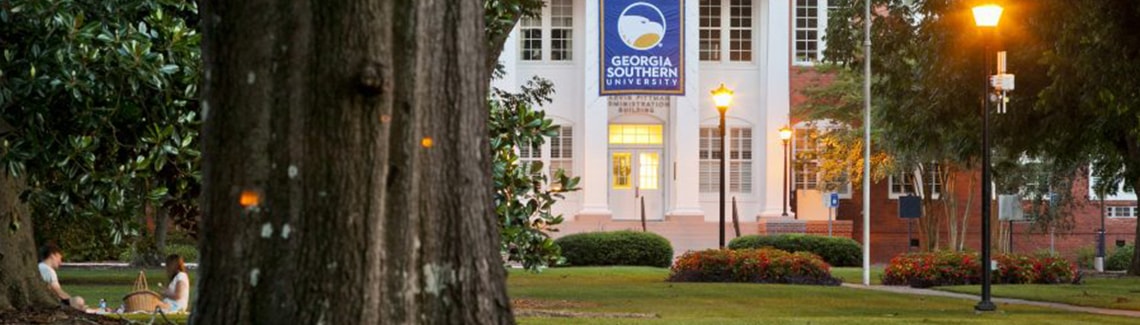 A banner-style photograph capturing the front entrance of the Marvin Pittman Administration Building at Georgia Southern University during dusk, featuring warm glowing outdoor lamps, a prominent university banner hanging between white columns, and a large tree trunk framing the left foreground.