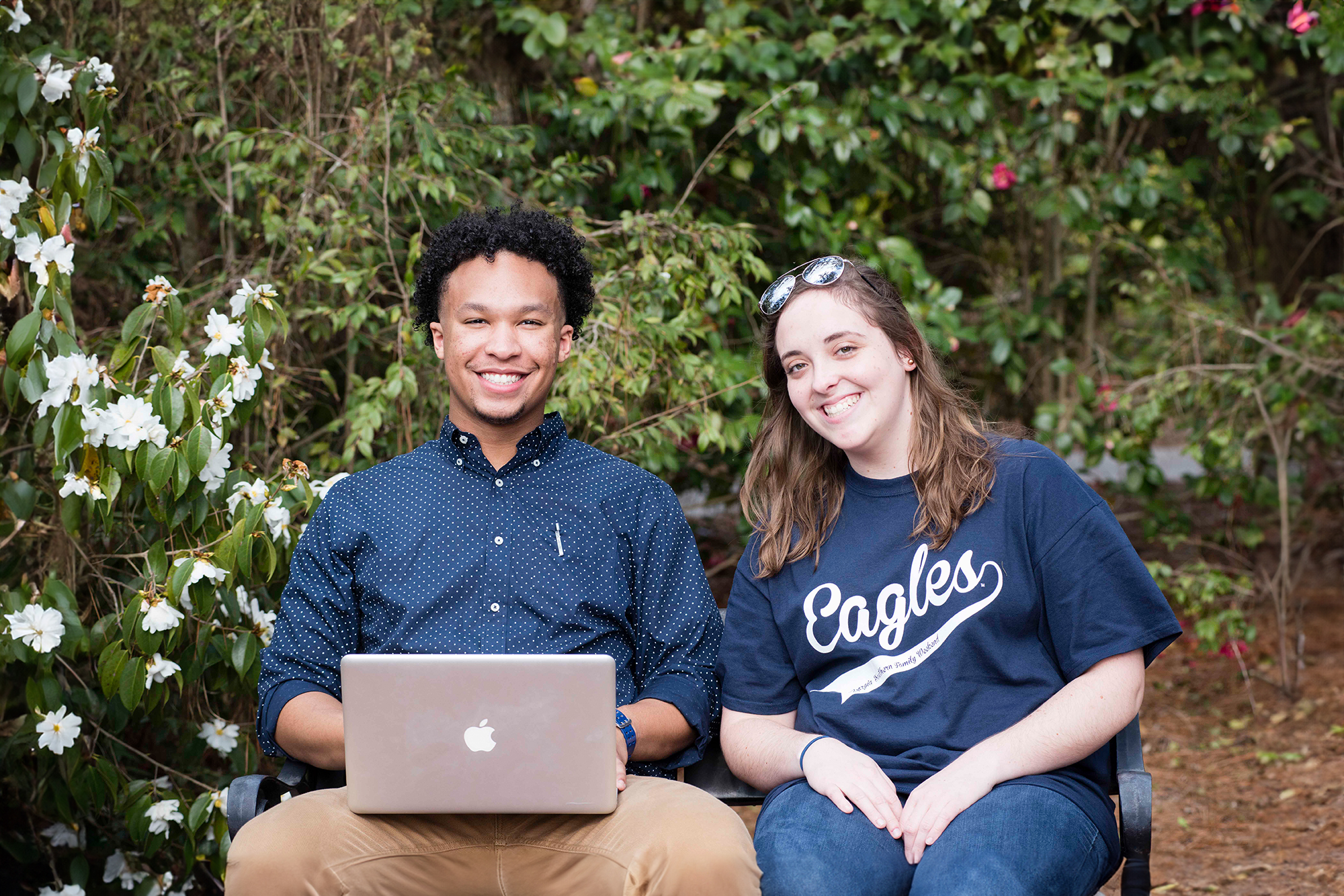 Two people sit on a bench outdoors in front of greenery and flowers. One person uses a laptop and smiles, while the other, wearing an Eagles shirt, sits beside them, also smiling.