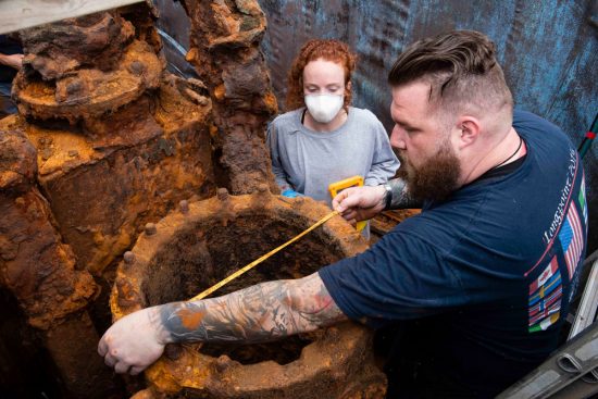 A male researcher with arm tattoos using a tape measure to take dimensions of a heavily rusted iron circular structure, observed by a colleague wearing a protective face mask.