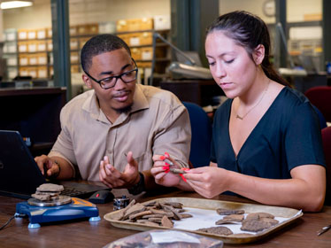 Two people sit at a table examining pottery shards; one holds and inspects a piece while the other gestures and looks on. A laptop and more artifacts are on the table in a well-lit workspace.
