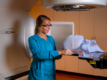 A healthcare professional in teal scrubs adjusts a white mesh mask on a patient positioning device under a large radiotherapy machine in a clinical setting.