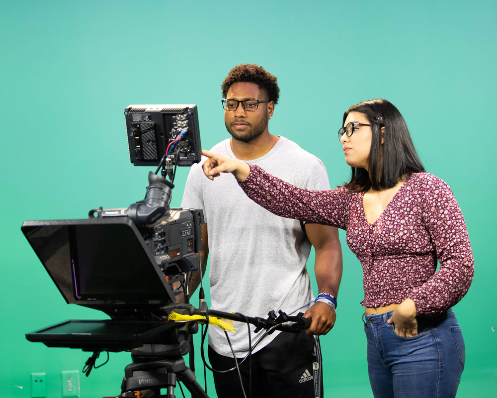 Two multimedia students operating a professional video camera and monitor in front of a green screen backdrop.