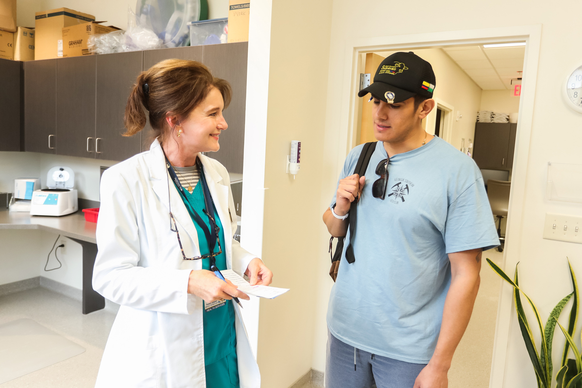 A female medical professional in a white lab coat talking to a young male student in a clinic doorway.
