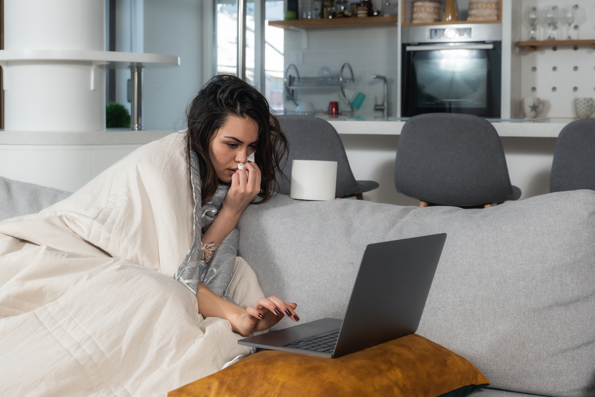 A sick woman wrapped in a blanket sitting on a couch and using a laptop while holding a tissue to her nose.