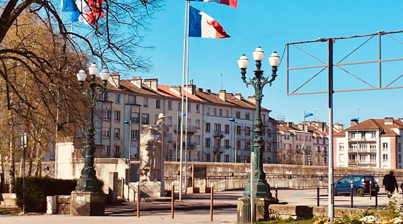 A street view of a public square in France featuring ornate green street lamps, a stone monument statue, and French flags waving on tall flagpoles in front of multi-story buildings under a clear blue sky.