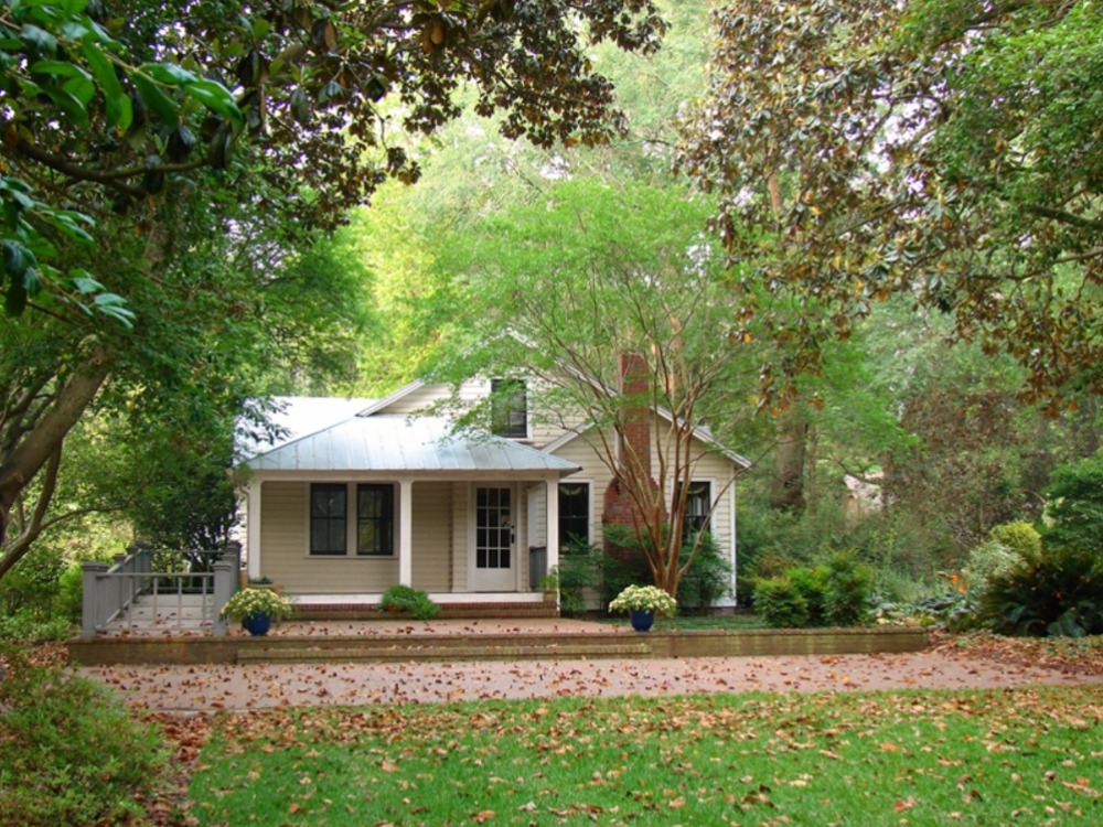 A small, light-colored cottage with a covered front porch and a brick driveway, nestled among mature green trees and lush landscaping with fallen autumn leaves on the ground.