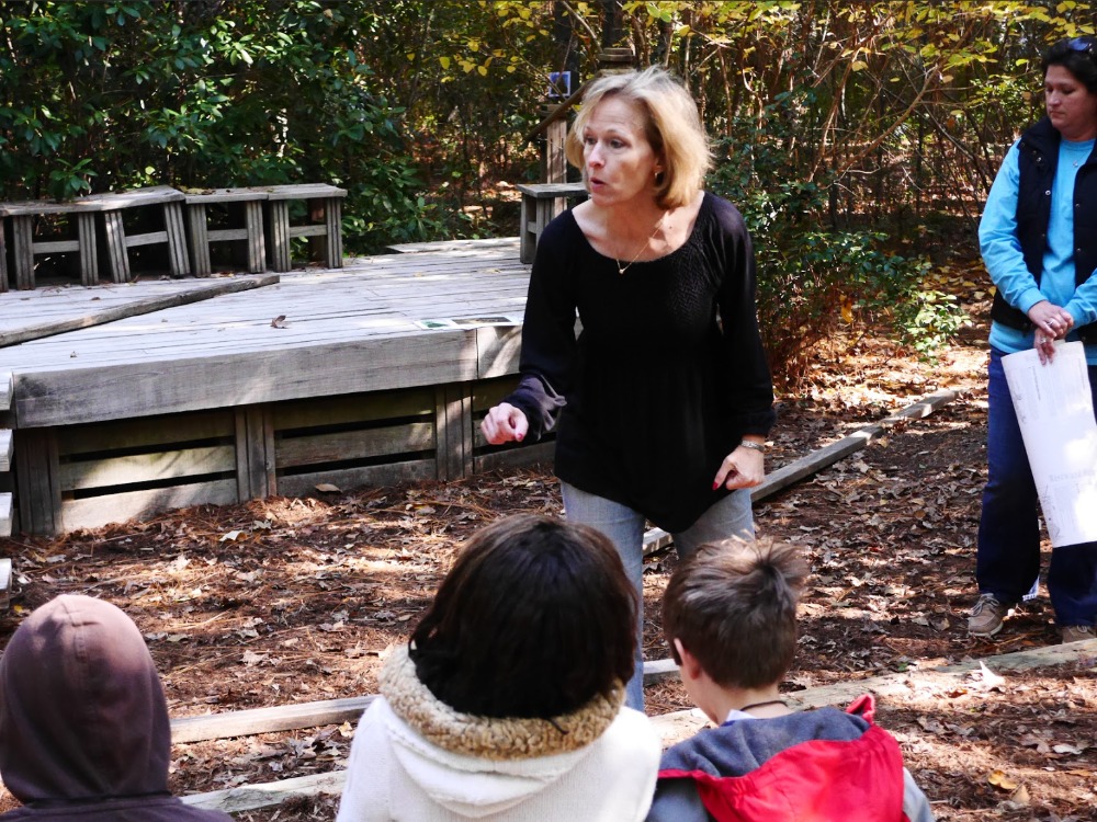 A woman in a black top gestures dynamically as she speaks to a group of children seated outdoors near a low wooden stage in a wooded area.