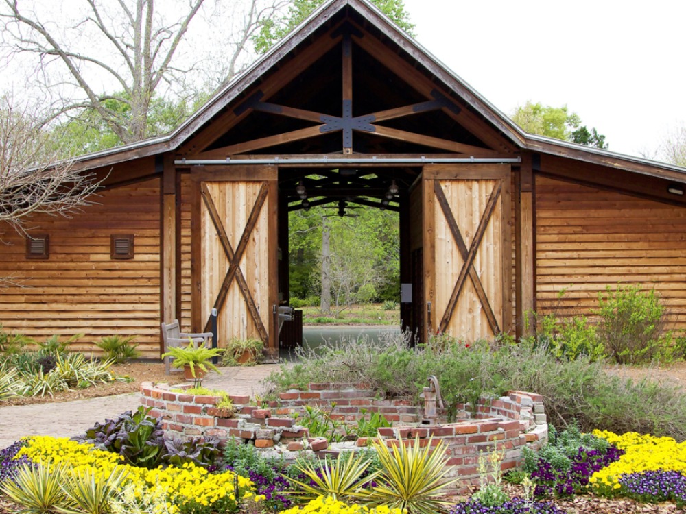 The exterior of a rustic wooden pavilion with large, open sliding barn doors that reveal a walkway through the building, fronted by a circular brick planter filled with flowers.
