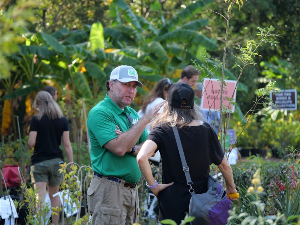 A plant nursery employee wearing a green polo shirt and a grey cap speaks with a customer in a busy outdoor garden center filled with plants and trees.