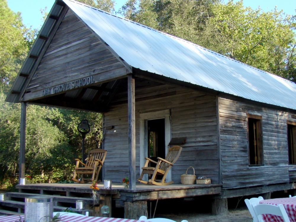 An old, weathered wooden building with a corrugated metal roof labeled "Oak Grove School," featuring a covered porch with two wooden rocking chairs.