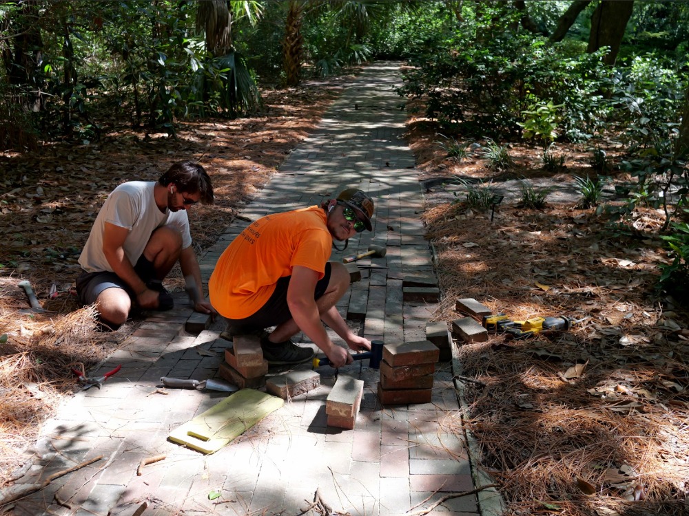 Two workers crouch on a brick walkway winding through a wooded garden area to repair and lay down paving bricks.