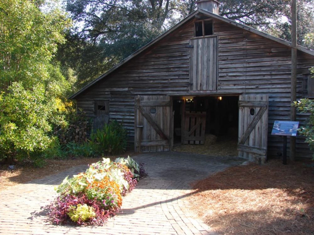A rustic, weathered wooden barn with its large doors wide open, facing a brick pathway lined with colorful flowerbeds and surrounded by lush green trees.