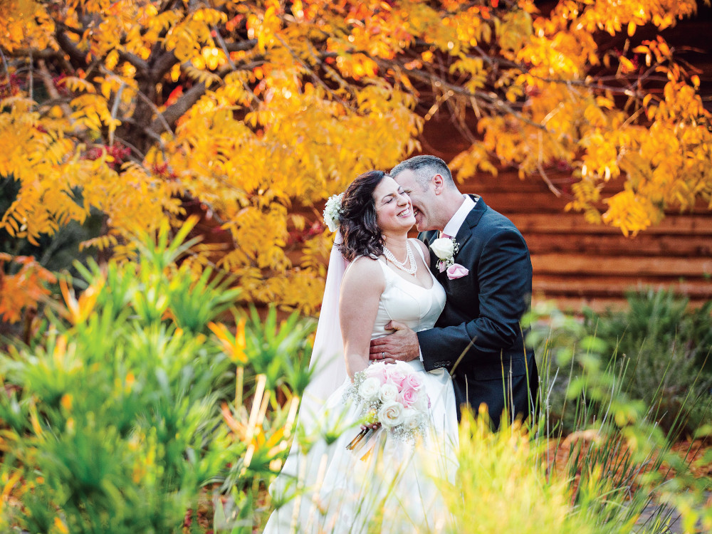 A joyful bride and groom laughing together outdoors in front of a tree with vibrant golden autumn foliage during their wedding day.