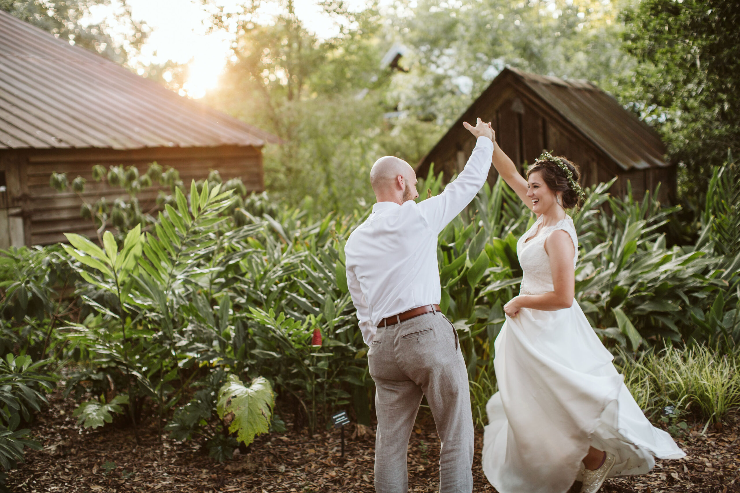 A happy bride and groom spinning and dancing together in an outdoor garden setting with rustic wooden sheds in the background during golden hour.