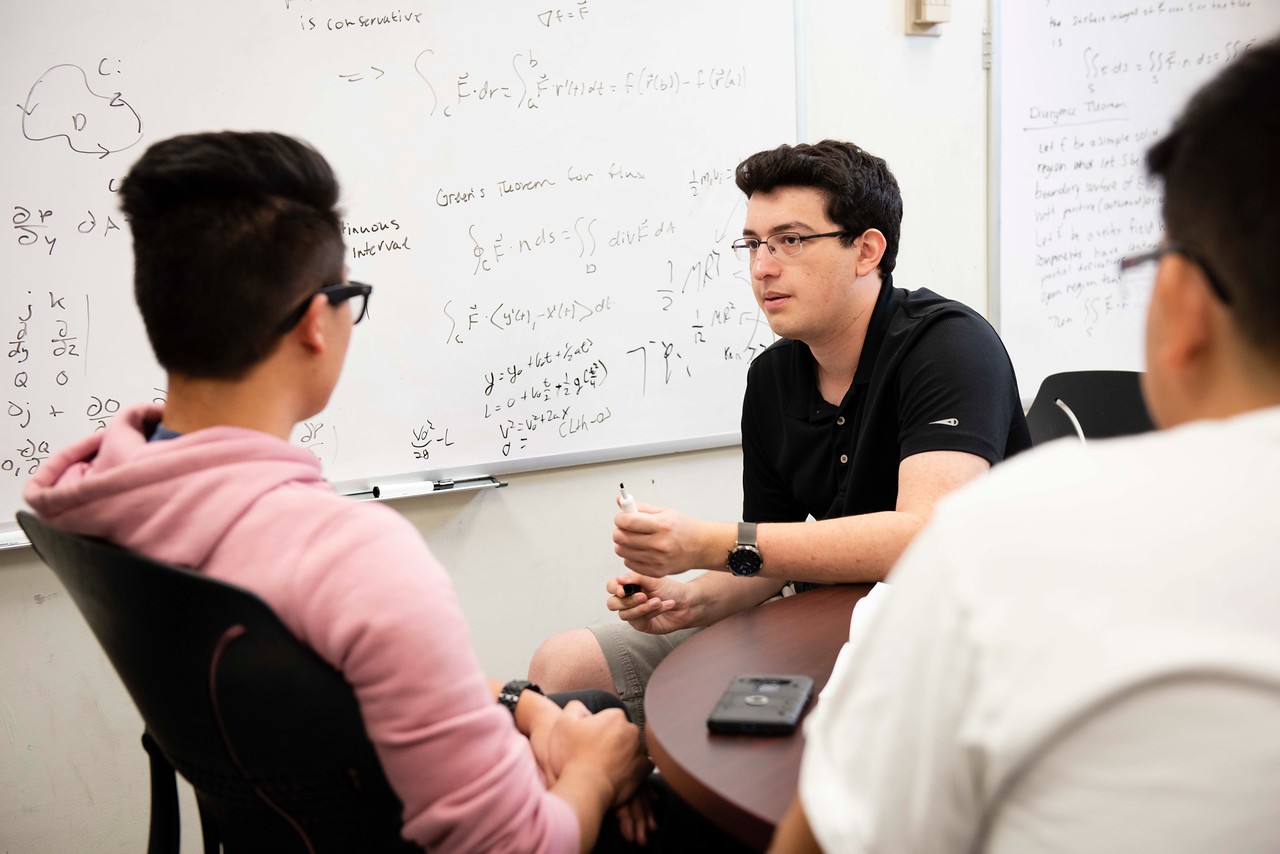 A male instructor or tutor wearing glasses and a black polo shirt holds a dry-erase marker while explaining complex math and physics formulas on a whiteboard to two students.