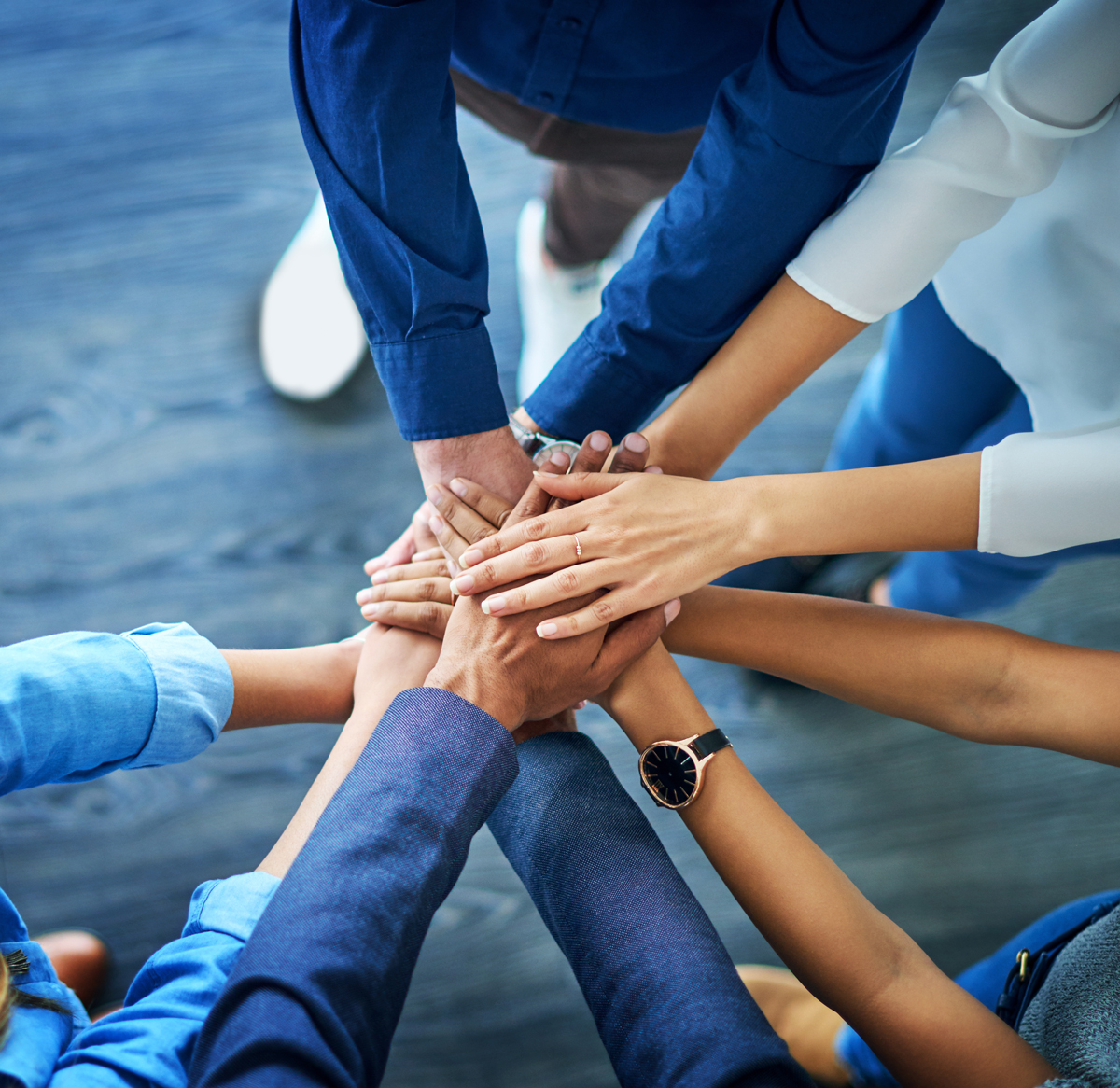 A top-down, close-up view of a diverse group of people stacking their hands together in the center of a circle to show unity and teamwork.