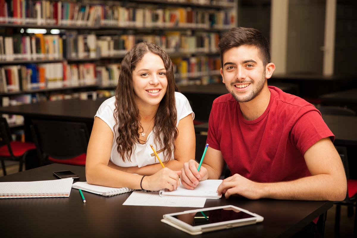 Two smiling students, a man and a woman, studying together with notebooks and pencils at a library table.
