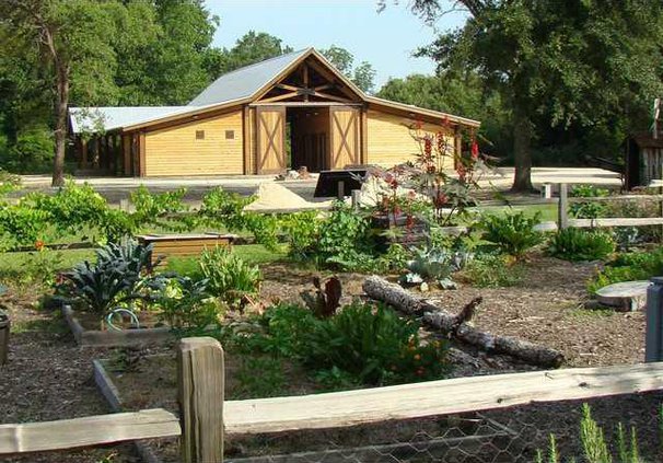 A scenic outdoor view of a fenced community garden with raised beds in the foreground, looking toward a large, rustic wooden barn style pavilion in the background.