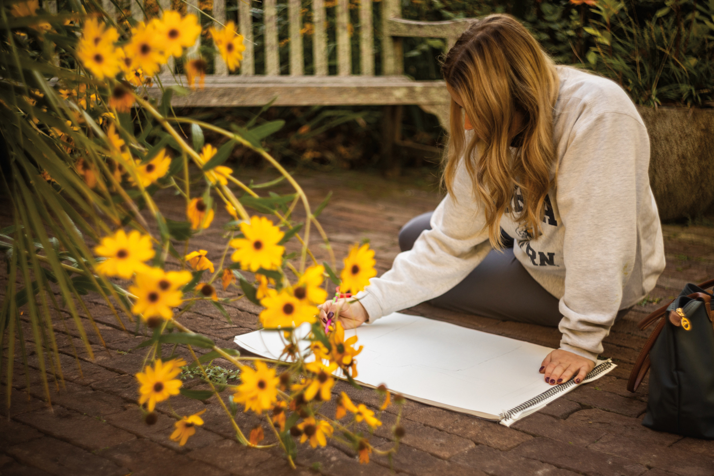A young woman kneels on a brick patio to sketch on a large sketchpad, with bright yellow black-eyed Susan flowers in the foreground.