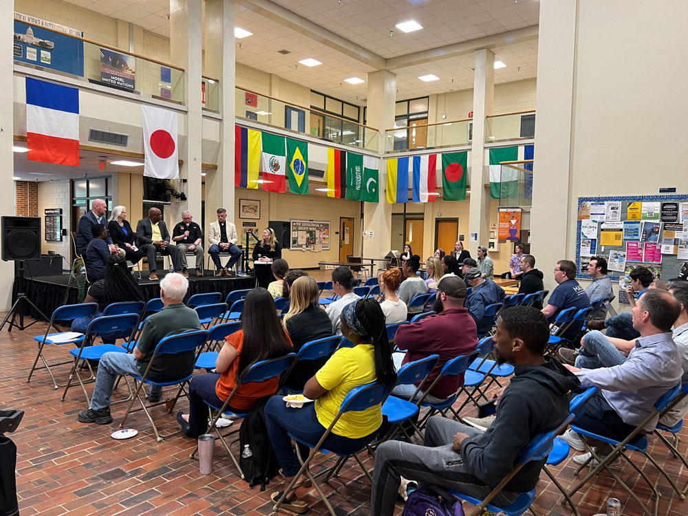 A panel of five speakers sits on a raised stage addressing an audience seated in blue folding chairs during an international-themed presentation inside a multi-story atrium decorated with world flags.