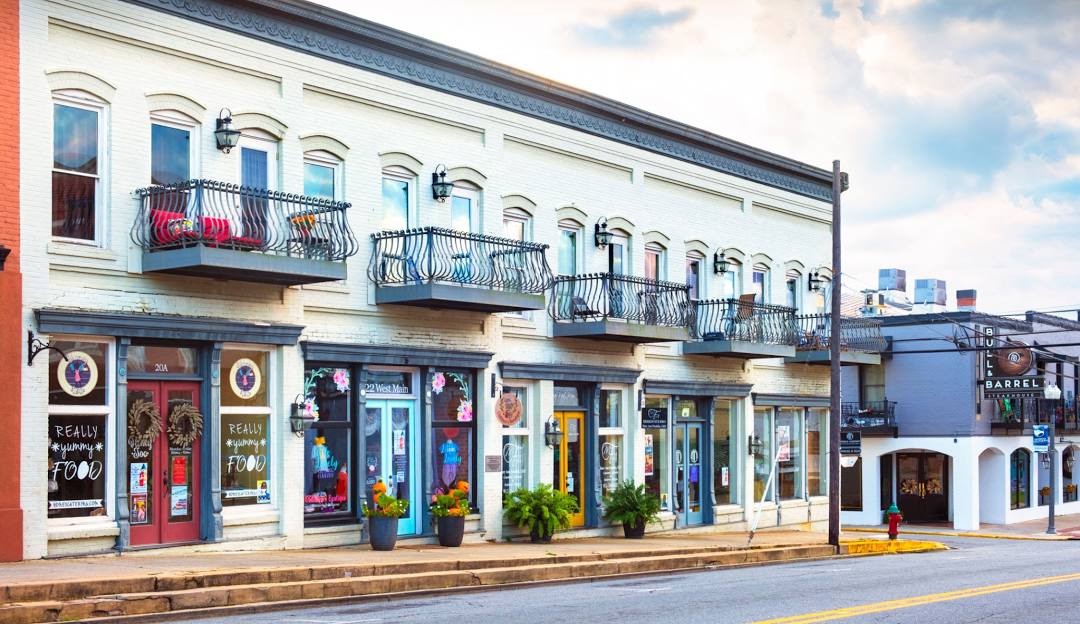 A row of historic two-story storefront buildings on a main street, featuring white brick facades, small balconies with black iron railings, and various local shops on the ground floor.