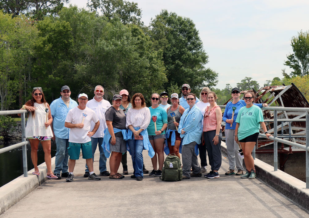 A large group of people stand together smiling on a concrete walkway outdoors near a body of water and a metal structure, mostly wearing casual shirts, shorts, and sunglasses.
