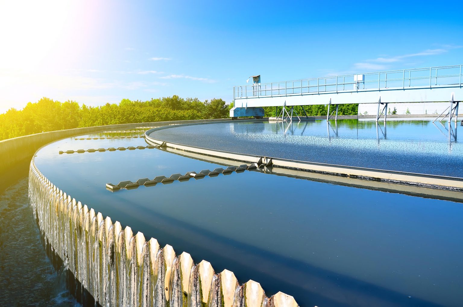 A wide outdoor shot of a large circular clarification tank at a water treatment facility, with water flowing over the weir edges under a bright, sunny blue sky.