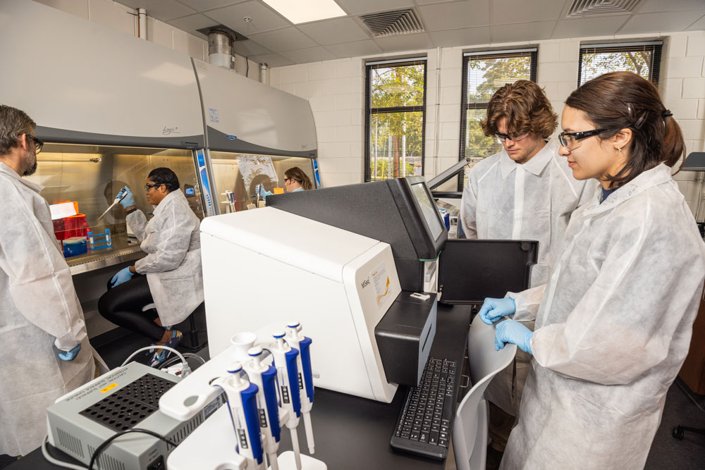 A team of scientists wearing lab coats, safety glasses, and blue gloves working in a modern laboratory equipped with biosafety cabinets and an analytical instrument on a counter.
