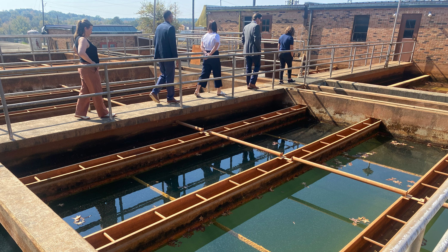 A group of five people walk along elevated concrete walkways with metal railings over open-air water treatment tanks containing clear water with fallen leaves.