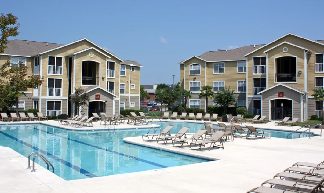 An outdoor swimming pool complex at an apartment community, featuring concrete decking lined with lounge chairs and surrounded by multi-story tan residential buildings under a clear sky.