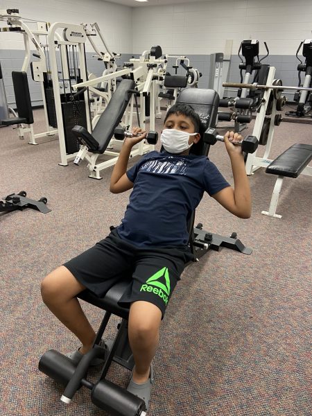 A young boy wearing a white face mask sits on an inclined weight bench while lifting small dumbbells in a gym.