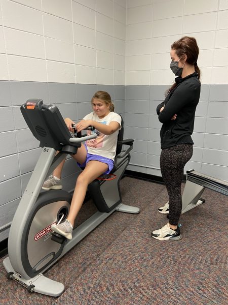 A woman wearing a face mask stands with her arms crossed while observing a young girl exercising on a recumbent stationary bike in a gym room.