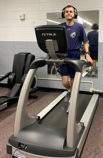 A young man wearing large headphones walks on a stationary treadmill while exercising in a gym room.