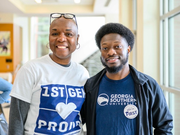 A close-up portrait of two smiling individuals standing side-by-side; the person on the left is wearing a 