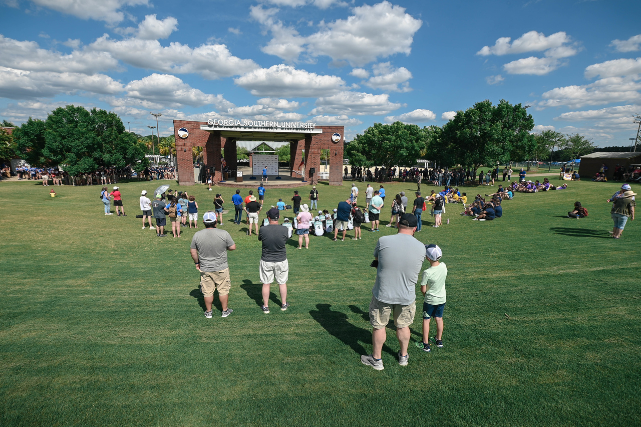 A high-angle view of a crowd gathered on a large grassy lawn watching an event at an outdoor stage with a sign that reads 