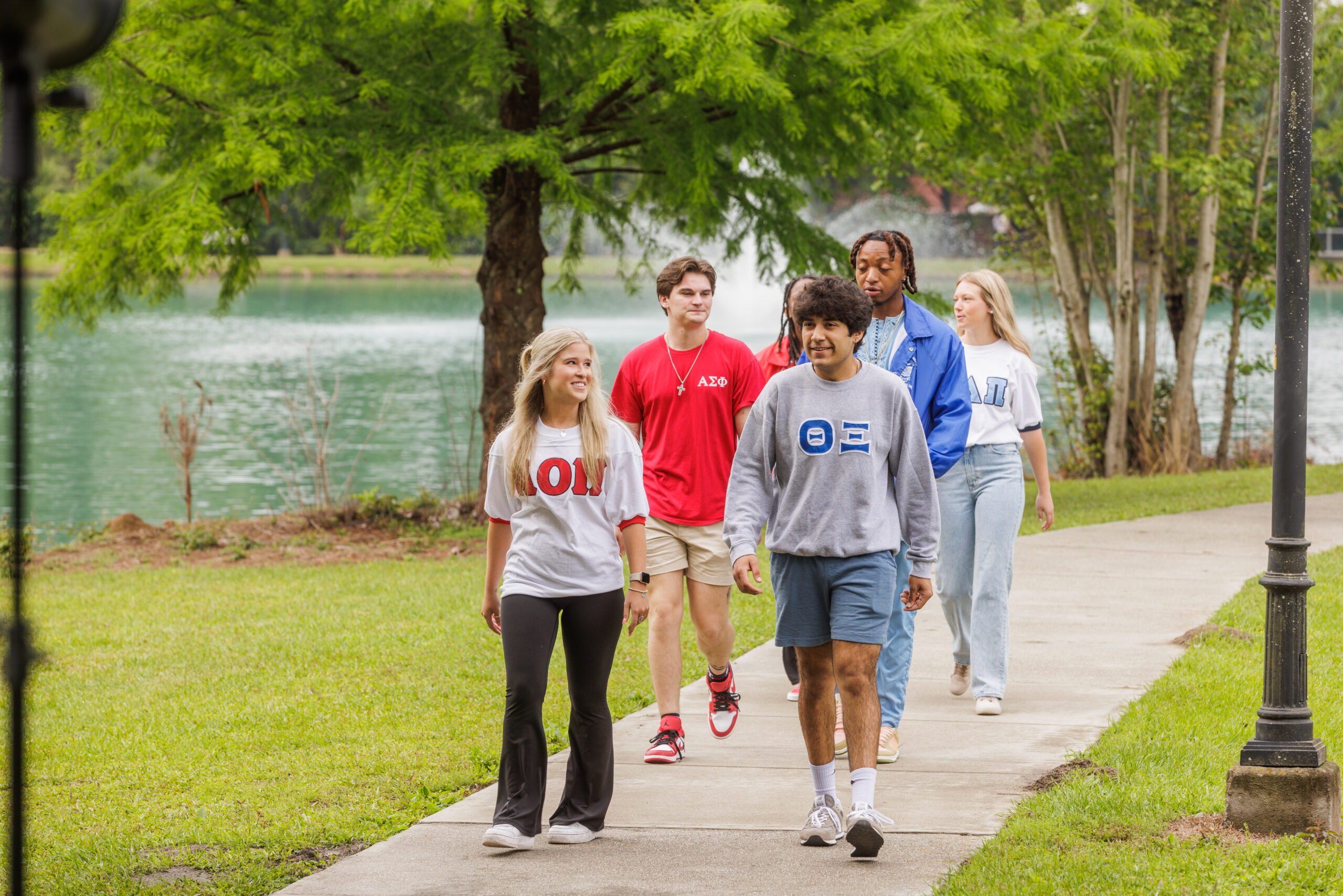 A group of university students wearing Greek life shirts and sweatshirts walk together along a paved campus pathway next to a lake with a fountain in the background.
