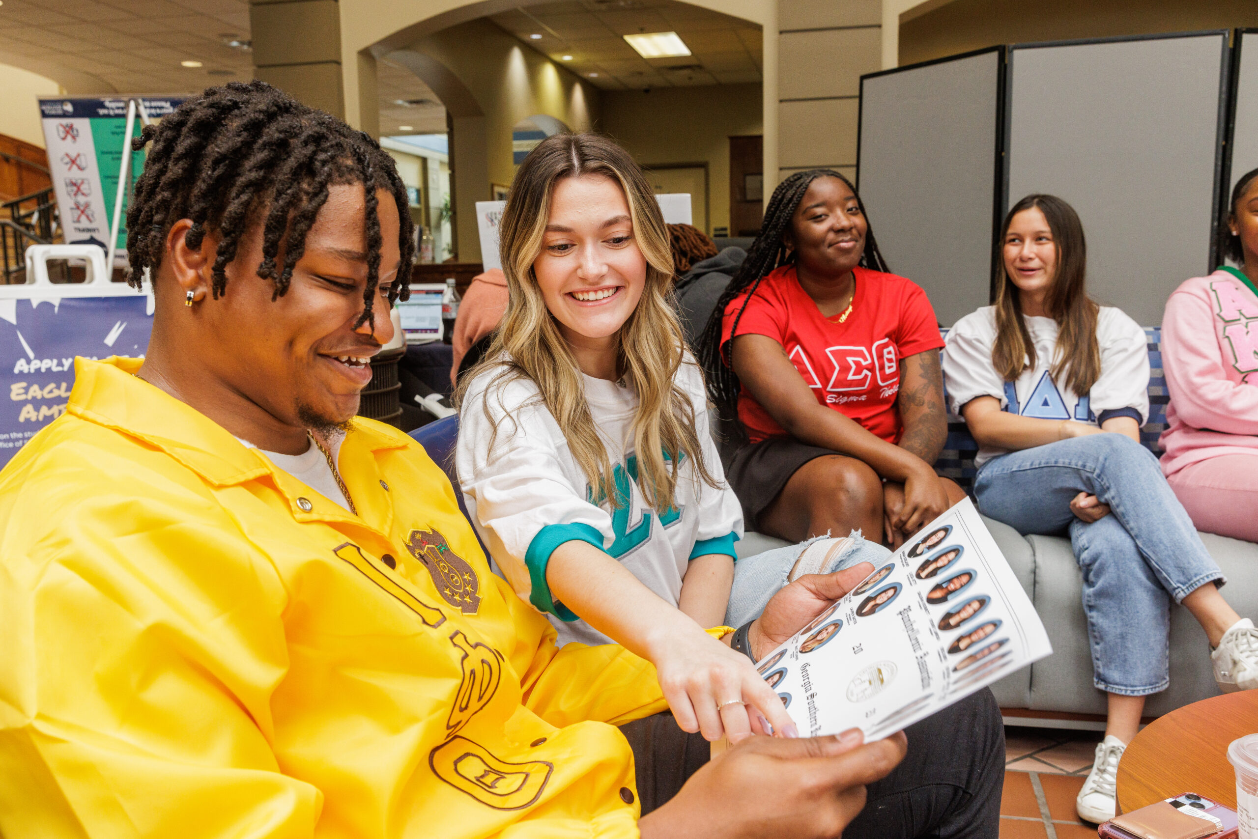 A group of university students sitting together indoors, with two students in the foreground smiling as they point at a printed composite sheet of fraternity or sorority member headshots.