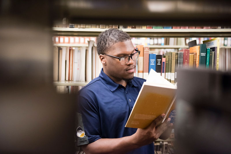 African American male Georgia Southern student standing in library isles looking at an open book in his hand.