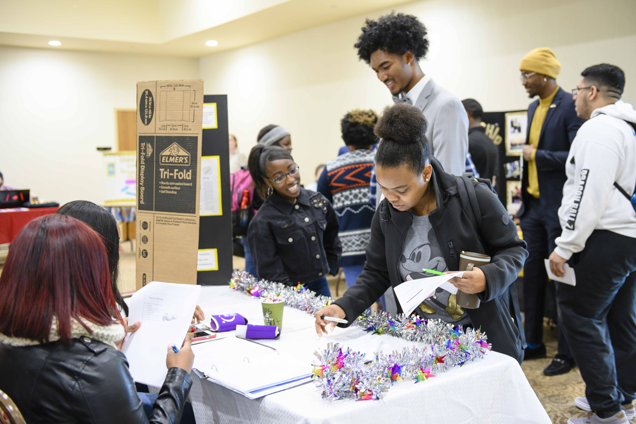 Students and visitors interact at an indoor campus event table decorated with tinsel garland, featuring tri-fold display boards in the background.